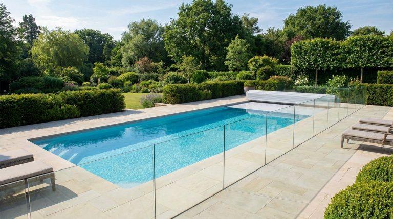 Piscine rectangulaire moderne avec eau bleue claire, entourée d'une élégante barrière en verre et d'une couverture automatique. Jardin verdoyant.