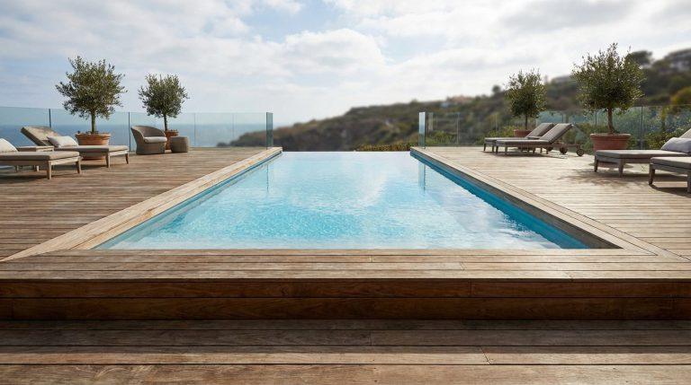Vue d'une piscine à débordement rectangulaire sur une grande terrasse en bois, avec des transats et des oliviers en pots.