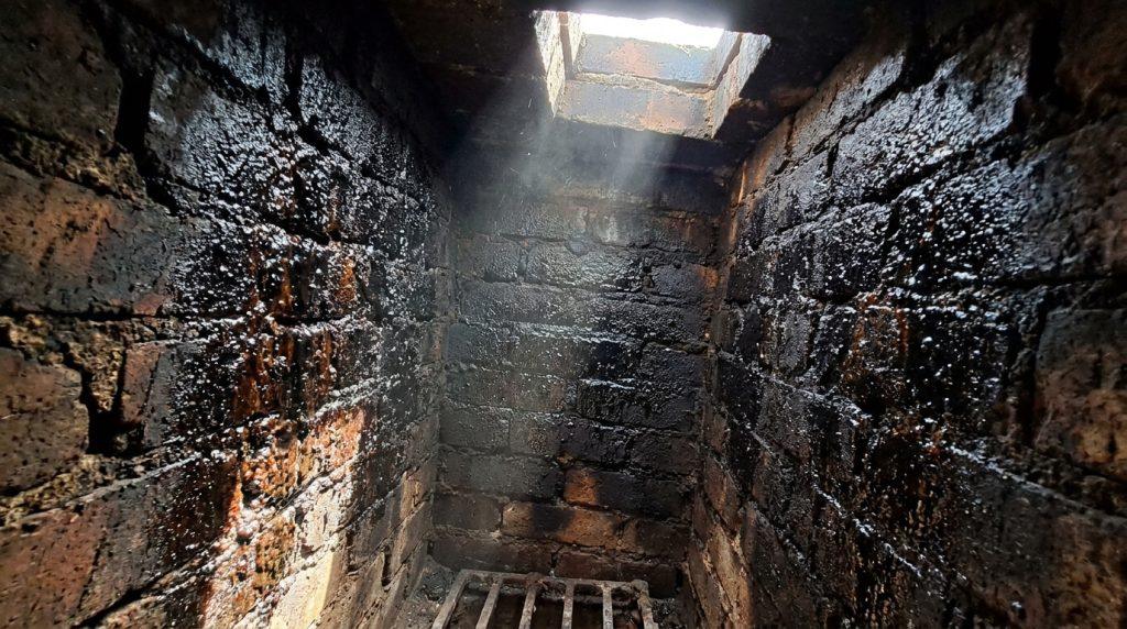 Intérieur de conduit de cheminée en briques, fortement encrassé de bistre noir et brillant. La lumière d'en haut révèle des dépôts.