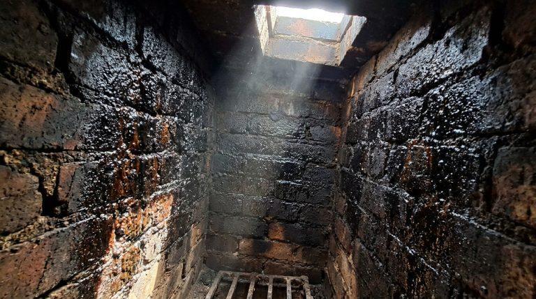 Intérieur de conduit de cheminée en briques, fortement encrassé de bistre noir et brillant. La lumière d'en haut révèle des dépôts.