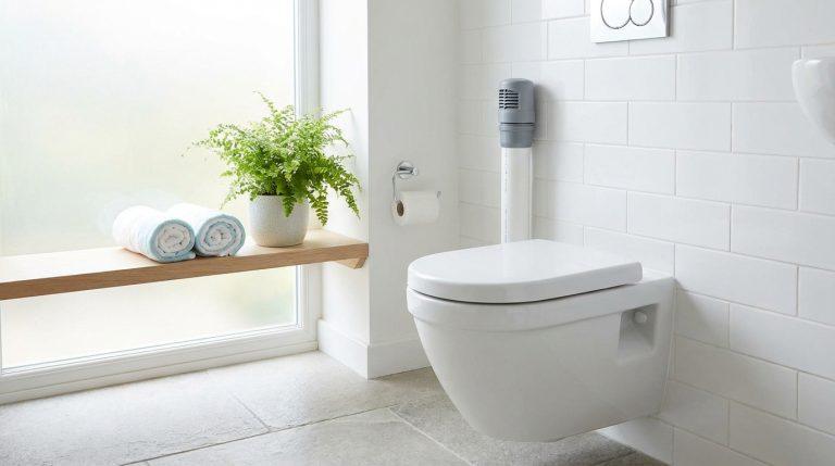 WC suspendu dans salle de bain moderne blanche. Système de décompression gris sur tuyau mural. Étagère bois, plante, serviettes et fenêtre dépolie.