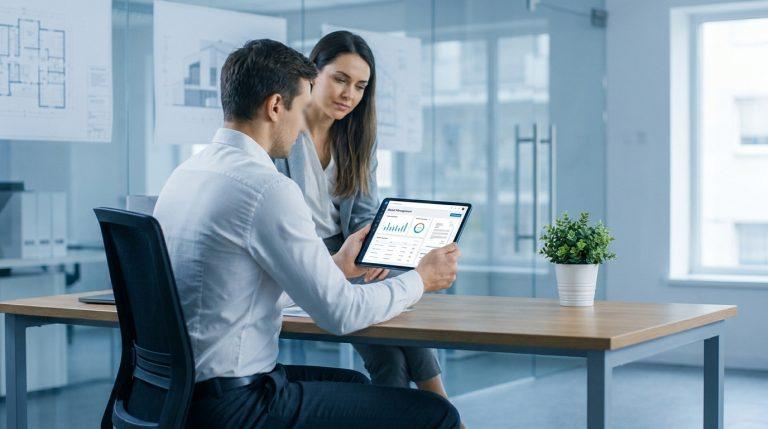 Un homme et une femme visualisent un tableau de bord de gestion locative sur une tablette, plans architecturaux en fond.